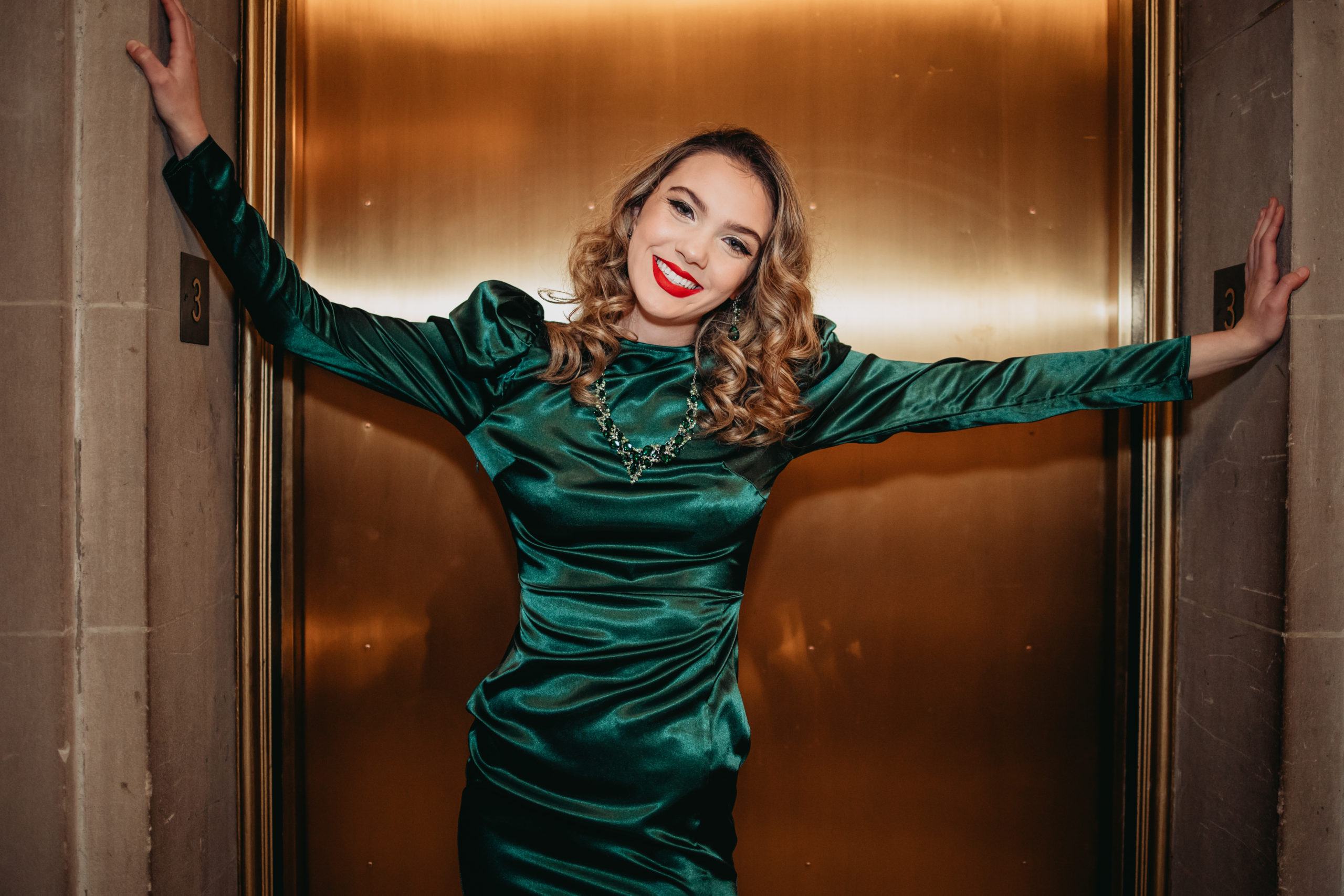 Glam Senior portraits at San Francisco City Hall.  Smiling girl dress in vintage style emerald green dress with beautiful red lipstick, standing in front of a shiny bronze elevator door.