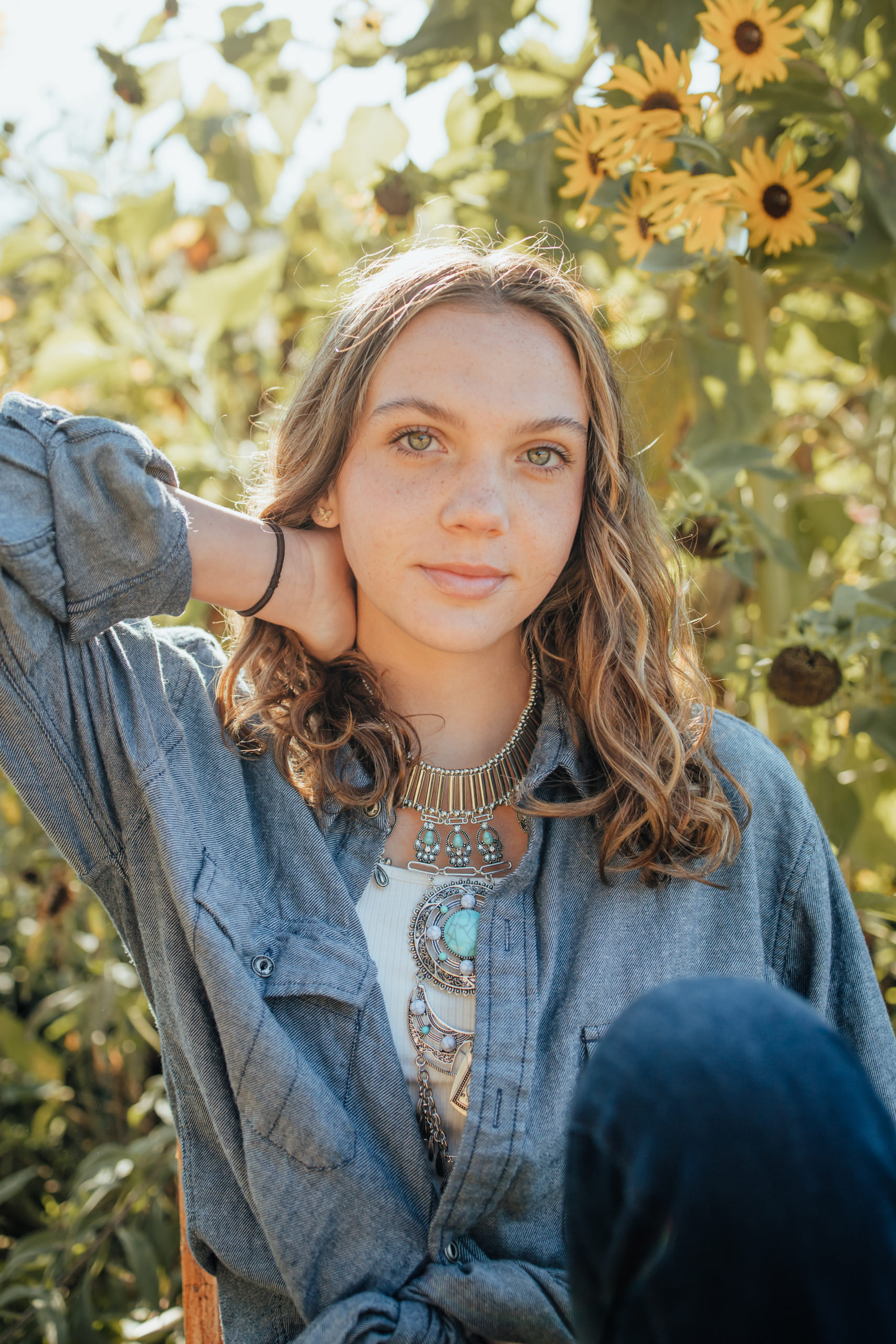 Senior portraits taken in Vacaville, at Soul Food Farm.  Girl with soft smile and freckles sitting in flower of sunflowers with denim shirt and turquoise necklace.