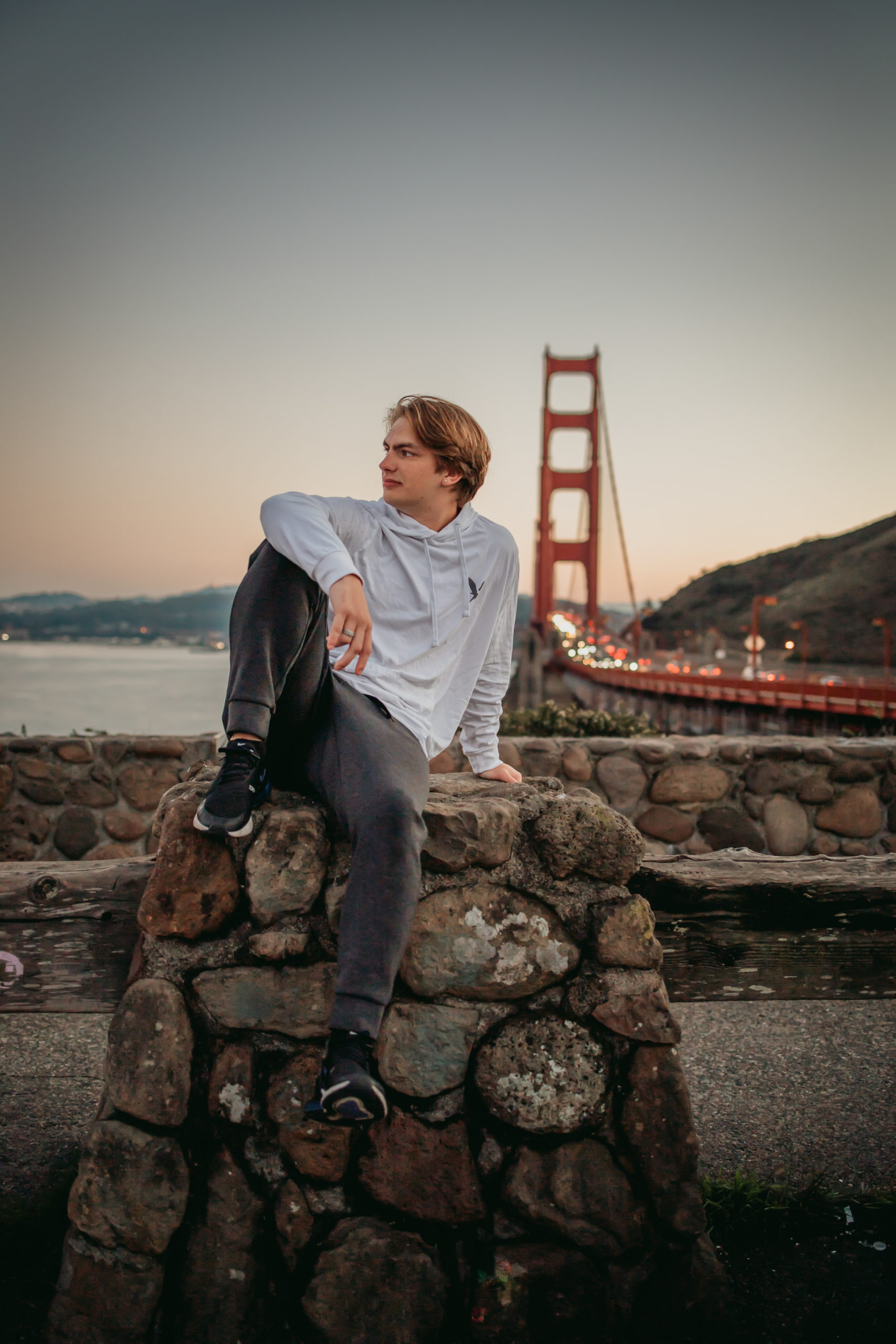 Senior portrait of boy who is causally dress.  Posing in front of the Golden Gate Bridge around dusk with headlights lit up on the bridge.